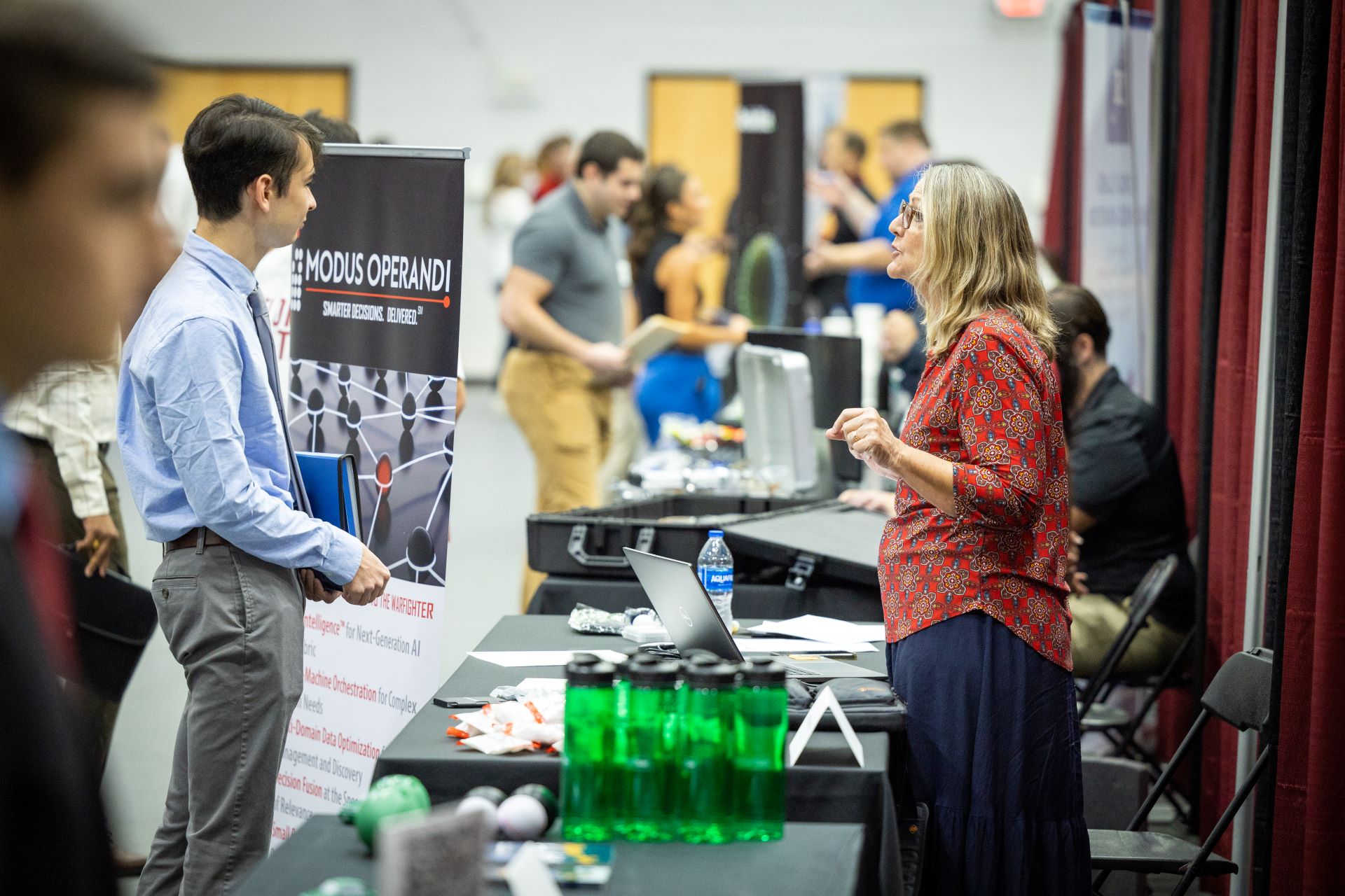 A career fair at Florida Tech where a young man in a light blue shirt speaks with a representative at a booth with a 'Modus Operandi' banner. Other attendees and booths are visible in the background